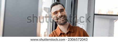 portrait of charismatic and joyful businessman in trendy eyeglasses and shirt smiling at camera while standing near grey wall in modern office, banner Royalty-Free Stock Photo #2306572043