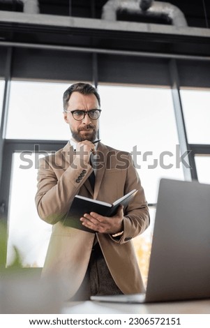 low angle view of thoughtful businessman in eyeglasses and stylish blazer standing with notebook and pen and looking at laptop in office on blurred foreground Royalty-Free Stock Photo #2306572151