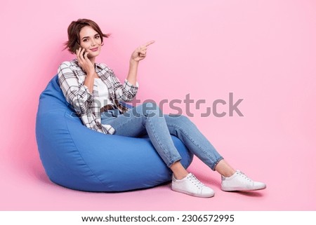 Full size photo of attractive girl sit comfort bag speak telephone direct finger empty space isolated on pink color background