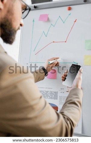 blurred bearded businessman in beige blazer holding smartphone with blank screen while drawing graphs of market research on flip chart in modern office