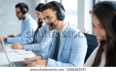 Confident handsome call center operator assisting customers, engaged in conversation, surrounded by colleagues in the Call Center office. Royalty-Free Stock Photo #2306583547
