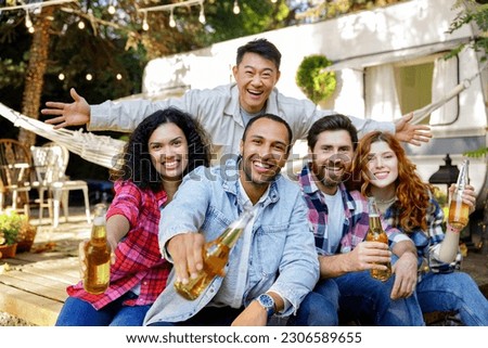 A cheerful and noisy company went on a picnic outside the city. Friends boys and girls of different races drinking drinks having fun near tourist trailer relaxing after work. Looking at the camera.