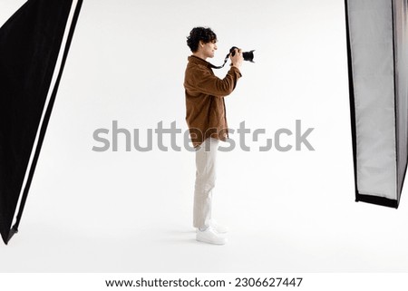 Full length shot of young photographer man holding professional photocamera, standing on white background with modern equipment, side view