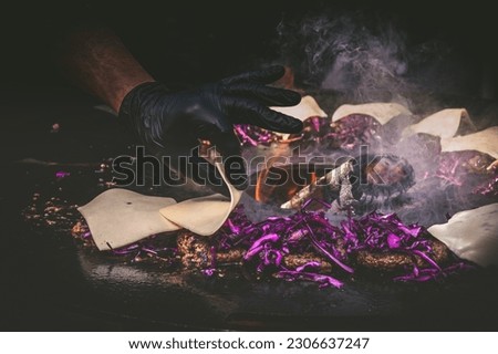Preparing juicy beef hamburger patties with red cabbage. BBQ meat sizzling over hot flames. A hand spreads cheese on the patties. Black background.