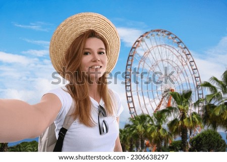 Beautiful woman in straw hat taking selfie near observation wheel outdoors