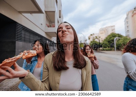 Young Caucasian woman with brown hair walking in front of friends and eating pizza joyfully.