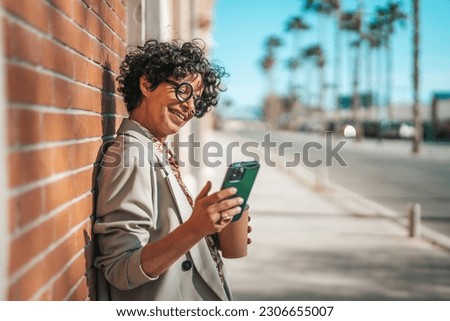 Waist up portrait of cheerful middle-aged lady in glasses talking on cellphone on the street. She is holding cup of coffee. Mature smiling woman in coat with coffee cup using mobile phone