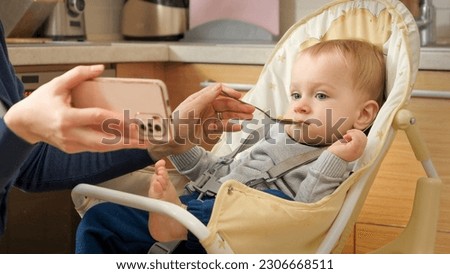 Portrait of little baby boy watching cartoons on smartphone while eating porridge in highchair. Concept of parenting, healthy nutrition and baby feeding