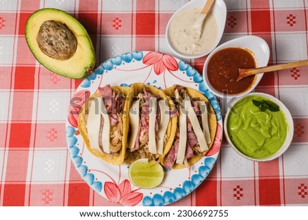 A top view of a plate of tacos next to dips and avocado, on a red checkered tablecloth