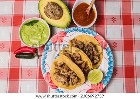 A top view of a plate of beef steak tacos next to dips and avocado, on a red checkered tablecloth