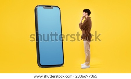 Excited Young Man Posing With Huge Cellphone Displaying Empty App Screen Standing On Yellow Studio Background. Guy Looking At Phone Touchscreen In Excitement, Advertising Mobile Offer. Panorama