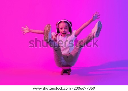 Childhood, kids hobbies and entertainment concept. Pretty emotional cute preteen stylish girl having fun on skateboard over studio background in neon light, using wireless headphones, copy space