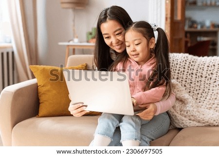 Family Digital Leisure. Happy Asian Mommy And Little Baby Daughter Using Laptop Learning And Having Fun Online Sitting On Couch At Home. Mother Showing Child Cartoons On Computer