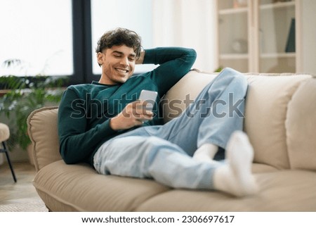 Mobile Offer. Smiling Middle Eastern Guy Browsing Internet On Smartphone, Communicating And Having Fun Online Lying On Couch At Home. Young Man Relaxing Using Gadget On Weekend. Selective Focus