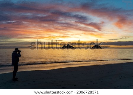 Photographer silhouette in pink, gold, and blue sunrise by calm ocean with Na Mokulua Twin Islands on the horizon