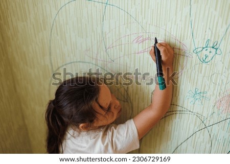 Top view of little naughty girl with dark hair wearing T-shirt, holding green marker in hand, drawing pictures on old yellow wallpaper. Art, misbehavior, redecoration, interior change, renovation.
