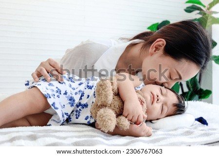 Cute toddle baby girl infant sleeping during hug teddy bear toy on bed, young beautiful mother kissing her sleepy daughter kid, mom comforting sleeping kid, parent take care child at home, love family