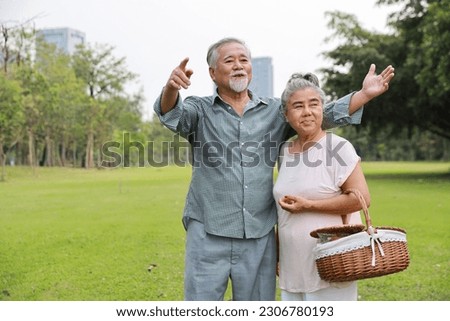 Happy asian senior man and woman walking and eyes contact while raising hands with picnic basket in garden outdoor. Lover couple going to picnic at the park. Happiness marriage lifestyle concept.