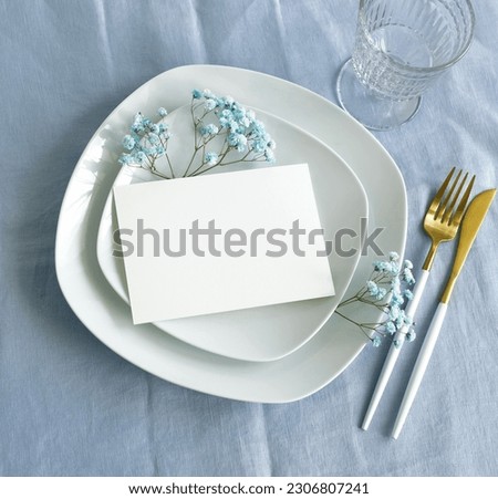 Card mockup on white plate with copy space and blue flowers top view on blue linen texture background .Invitation, menu. Blank paper, sheet card mock up.