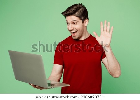 Young happy smart IT man he wearing red t-shirt casual clothes hold use work on laptop pc computer waving hand get video call isolated on plain pastel light green background studio. Lifestyle concept