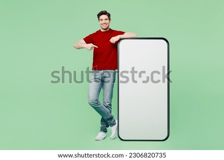 Full body young man he wearing red t-shirt casual clothes point index finger on big huge blank screen mobile cell phone smartphone with workspace area isolated on plain pastel light green background
