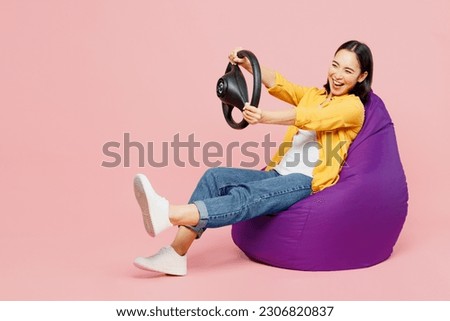 Full body fun young woman of Asian ethnicity wear yellow shirt white t-shirt sit in bag chair hold steering wheel driving car isolated on plain pastel light pink background studio. Lifestyle concept Royalty-Free Stock Photo #2306820837