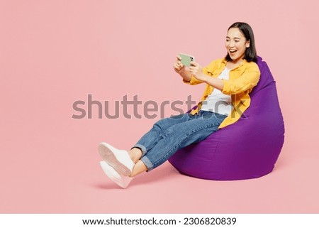 Full body young woman of Asian ethnicity in yellow shirt white t-shirt sit in bag chair use play racing app on mobile cell phone gadget smartphone for pc video games isolated on plain pink background
