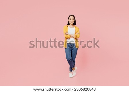 Full body smiling cheerful fun cool young woman of Asian ethnicity wear yellow shirt white t-shirt hold hands crossed folded look camera isolated on plain pastel light pink background studio portrait
