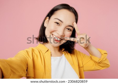 Close up young smiling woman of Asian ethnicity wear yellow shirt white t-shirt doing selfie shot pov on mobile cell phone show v-sign isolated on plain pastel light pink background studio portrait
