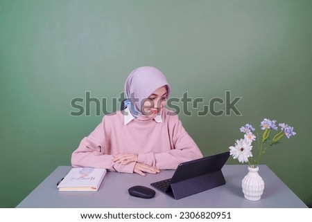 a Muslim woman wearing a hijab is at a desk watching her tablet in front of a green background