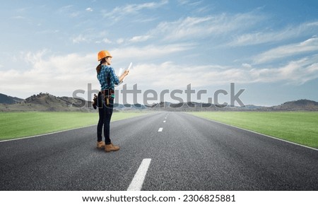 Young woman in safety helmet standing on asphalt highway with blueprint. Back view architect in workwear checking construction on nature background. Road infrastructure reconstruction and development