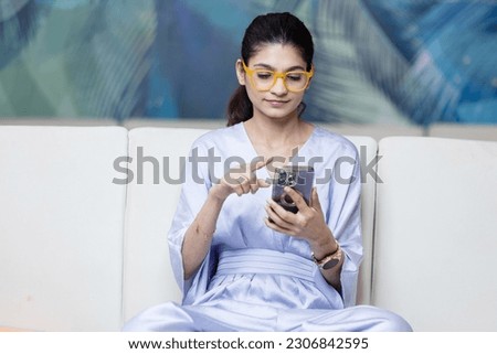 Happy indian lady relax at home alone sit on large couch in comfortable pose share good news at social media via cellphone. 