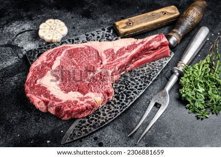 Dry aged raw tomahawk or cowboy beef steak on a black table with knife, cleaver, spice and herbs. Black background. Top view.