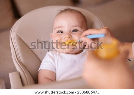 Cheerful baby boy being fed with porridge while sitting in high chair all messy and staied smiling and eating Royalty-Free Stock Photo #2306886475