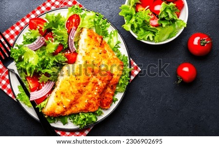 Fried white fish fillet with vegetable salad from lettuce, cherry tomatoes and red onion, black table background, top view