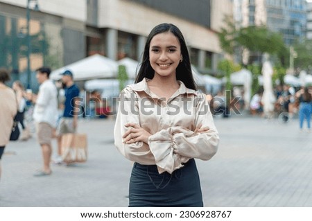A pretty asian woman standing outside wearing a light tan silk blouse and a high waist skirt, arms crossed while smiling prettily at the camera. A building, people and tents in the background