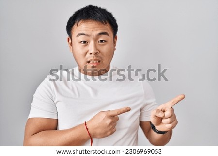 Young chinese man standing over white background pointing aside worried and nervous with both hands, concerned and surprised expression 