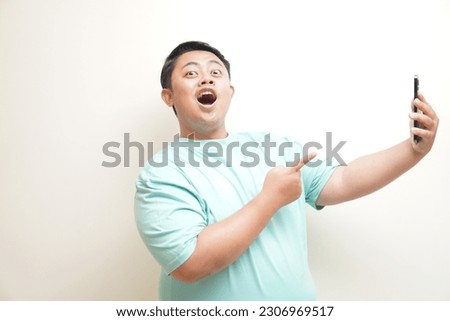 Portrait of attractive Asian man in green t-shirt holding mobile phone with excited expression. Isolated image on white background