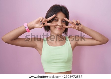 Young girl standing over pink background doing peace symbol with fingers over face, smiling cheerful showing victory 