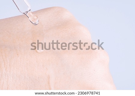 Macro photo of woman's hand and pipette with gel on blue background. Transparent drop of essence or gel drops from the pipette on the  extra dry skin, close up. SPA concept. Royalty-Free Stock Photo #2306978741