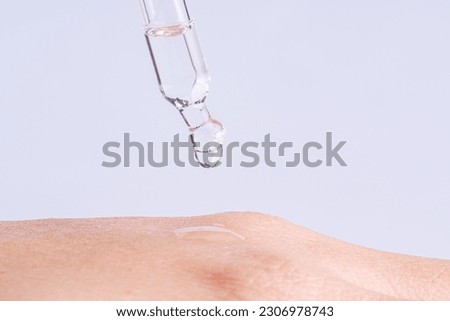 Macro photo of woman's hand and pipette with gel on blue background. Transparent drop of essence or gel drops from the pipette on the  extra dry skin, close up. SPA concept. Royalty-Free Stock Photo #2306978743