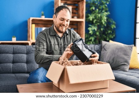 Young hispanic man unpacking virtual reality glasses sitting on sofa at home