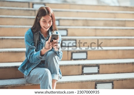 Happy young asian woman sitting on steps outdoors using social media on mobile phone. Royalty-Free Stock Photo #2307000935