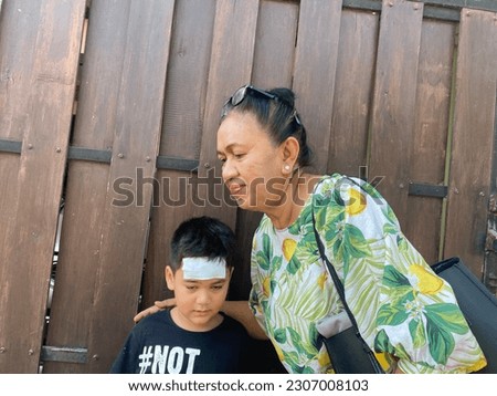 Grandma wearing floral blouse and grandson wearing black tee shirt writing not bother and compress on forehead, taking picture together front of house gate