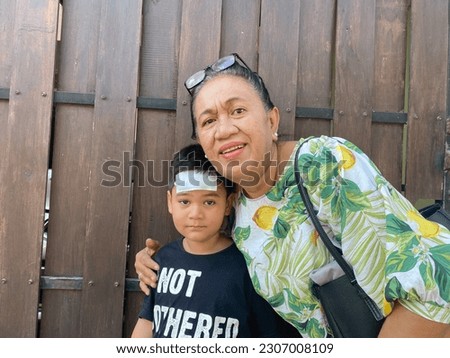 Grandma wearing floral blouse and grandson wearing black tee shirt writing not bother and compress on forehead, taking picture together front of house gate
