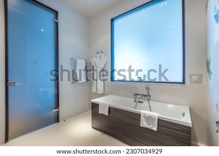 Stylish design of the bathroom in the hotel, with a frosted glass door and a false window. Bath with shower, heated towel rail and bathrobes. Royalty-Free Stock Photo #2307034929
