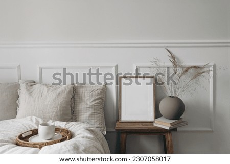 Elegant boho bedroom interior. Vertical wooden picture frame mockup on old bedside table. Cup of coffee. Ball vase with dry grass. Beige linen throw bedding. White wall background. Scandinavian home.