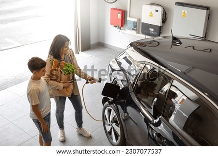 Happy son and mother plug EV charger from home charging station to electric vehicle. Future eco-friendly car powered by renewable source of clean energy on daytime. Horizontal, high angle. Royalty-Free Stock Photo #2307077537