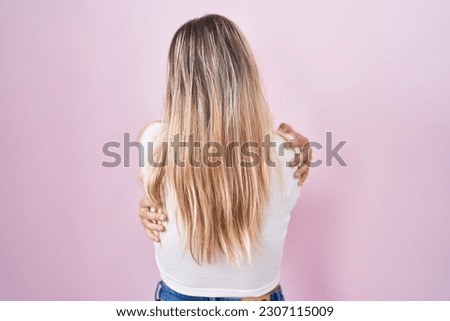 Young blonde woman standing over pink background hugging oneself happy and positive from backwards. self love and self care 