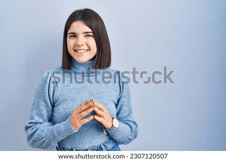 Young hispanic woman standing over blue background hands together and fingers crossed smiling relaxed and cheerful. success and optimistic 